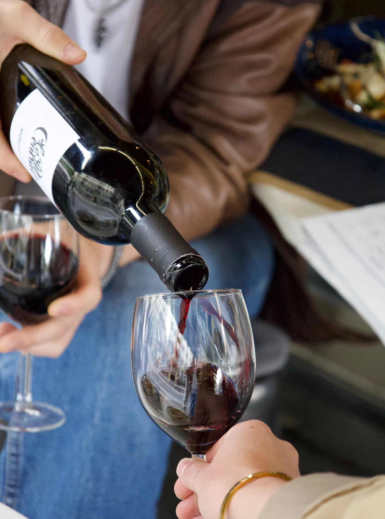 Le merlot de la maison versé de sa bouteille dans un verre tenu à table ; un second verre, déjà servi, attend à côté.